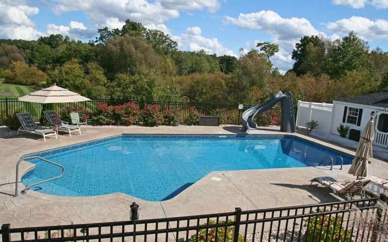 This is a photo of a true Lazy L style custom inground swimming pool with a black fence, steps and water slide.